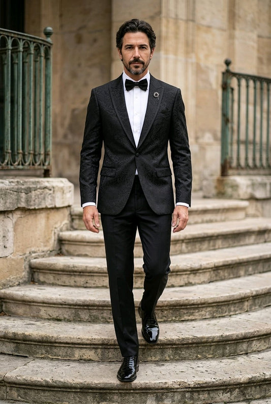 A man wearing a black textured tuxedo jacket with matching trousers, a white dress shirt, and a black bow tie walks down stone steps in front of a historic stone building.