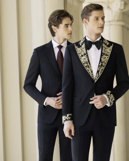 A man wearing a black tuxedo with gold brocade detailing on the lapels, single-breasted button fastening, and black bow tie, standing in a columned hallway.