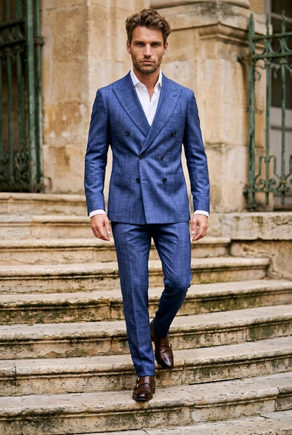 A man wearing a blue double-breasted suit with a subtle check pattern, a white dress shirt, and brown leather shoes, walking down stone steps in front of a historic building.