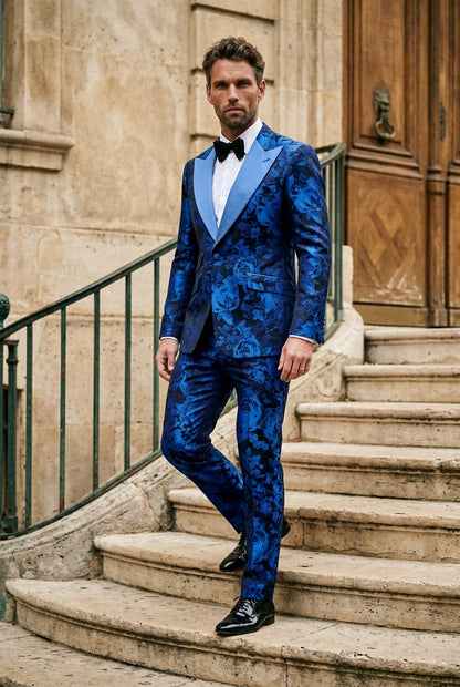 A man wearing a blue floral-patterned tuxedo with satin lapels, a white shirt, and a black bow tie, posing on stone stairs in front of a historic building.