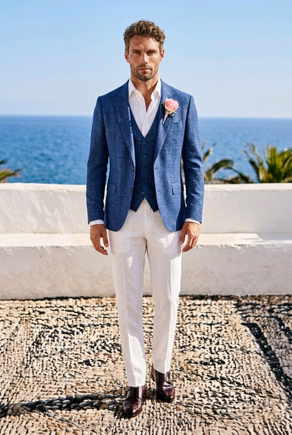 A man wearing a blue three-piece suit with white trousers and a pink boutonniere, standing outdoors on a stone path with the ocean and white wall in the background.