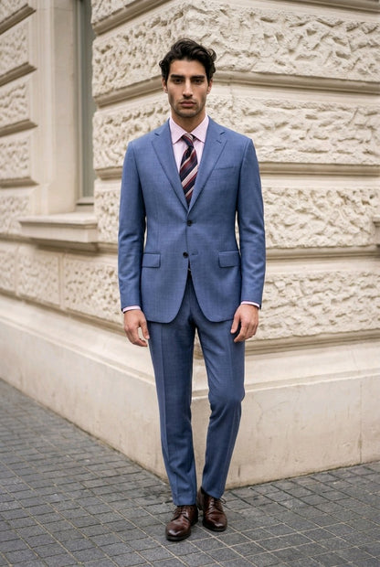 A man wearing a blue two-piece suit with a light pink dress shirt and a striped tie, standing outdoors against a textured beige stone wall.