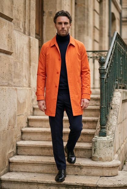 A man wearing a bright orange trench coat over a black turtleneck sweater, dark navy trousers, and black leather shoes, walking down stone stairs in an urban setting.