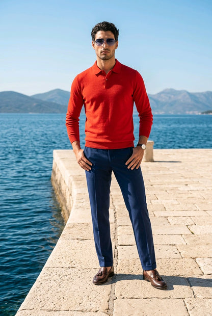 A man wearing a bright red long-sleeve polo sweater, navy blue trousers, and brown leather loafers stands on a stone pier with a sea and mountains in the background.