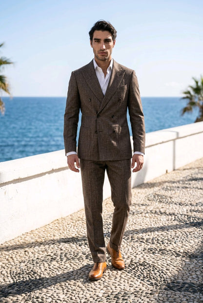 A man wearing a brown double-breasted plaid suit with a white shirt and brown leather shoes, standing on a cobblestone path with the blue ocean and sky in the background.