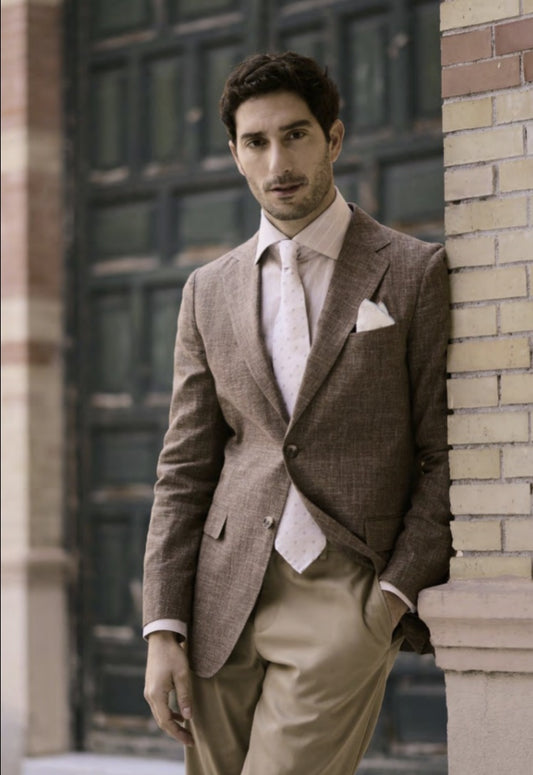 A man wearing a brown linen & cotton jacket with beige chino trousers, standing against a brick wall backdrop.