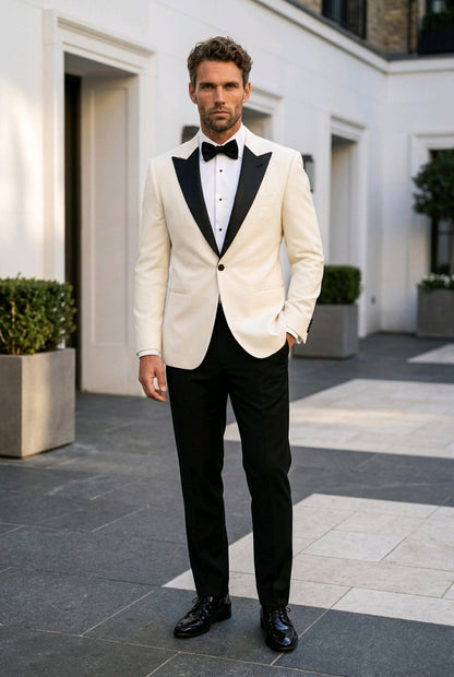 A man wearing a cream-colored tuxedo jacket with black satin lapels, black trousers, a black bow tie, and a white dress shirt, standing in an outdoor courtyard setting.