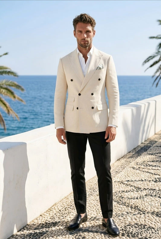 A man wearing a cream double-breasted blazer with black trousers and black dress shoes, standing on a stone path with the blue sea and sky in the background.