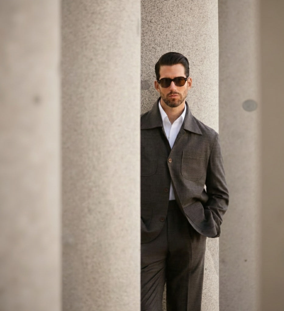 A man wearing a dark brown two piece suit with sunglasses, standing against a column.