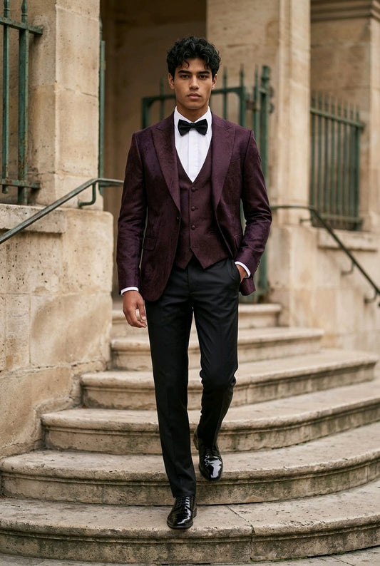 A man wearing a dark burgundy tuxedo jacket and matching vest with black trousers, a white shirt, and a black bow tie walks down stone steps in front of a classical building.