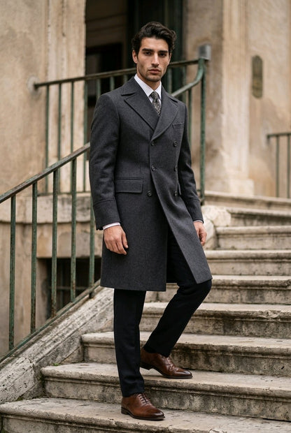 A man wearing a dark charcoal gray double-breasted overcoat, black trousers, a white shirt, and brown leather dress shoes, standing on stone stairs in front of a building.