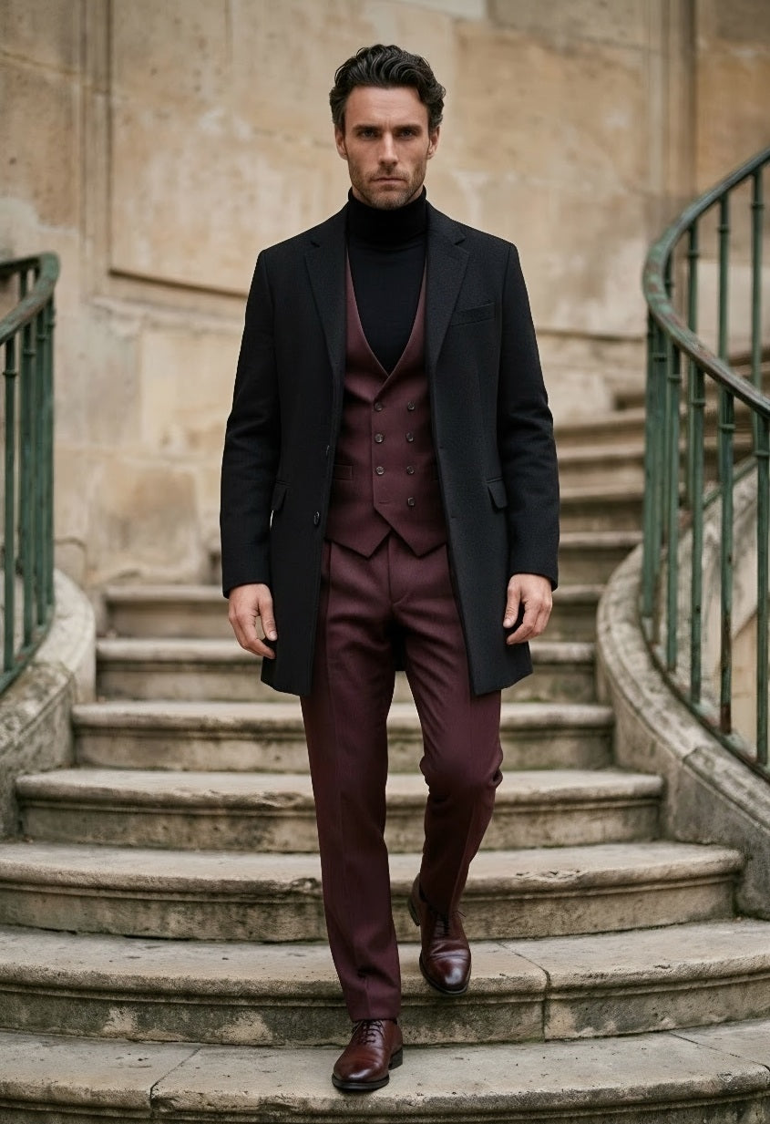 A man wearing a dark charcoal overcoat, burgundy three-piece suit, black turtleneck, and brown leather dress shoes walks down stone stairs in an outdoor setting with stone walls and green railings.