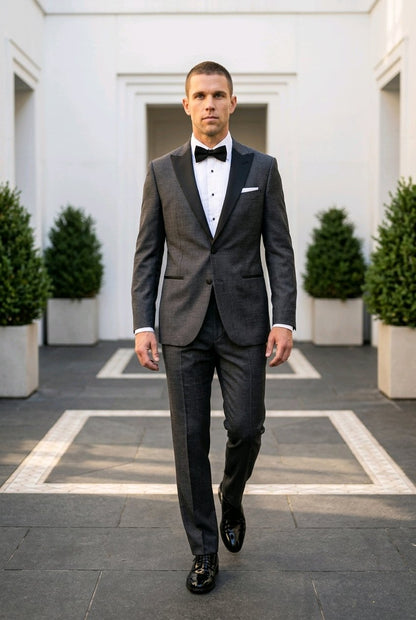 A man wearing a dark gray textured tuxedo with black satin lapels, a white dress shirt, and a black bow tie walks toward the camera in an elegant outdoor courtyard with white walls and potted greenery.