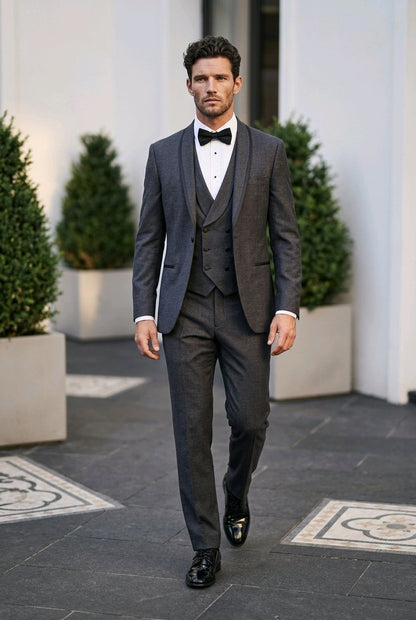 A man wearing a dark gray three-piece tuxedo with a white shirt, black bow tie, and black dress shoes, walking outdoors on a paved path with potted green shrubs.