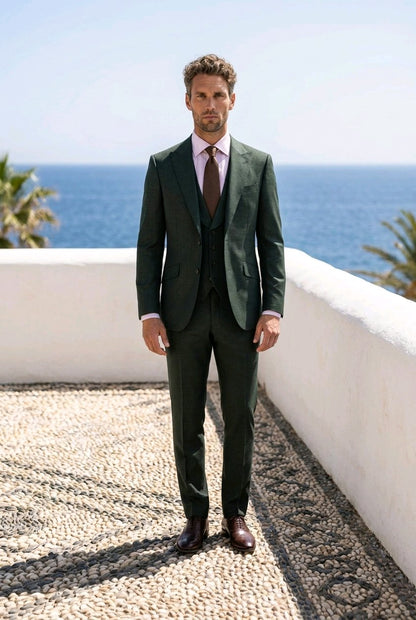 A man wearing a dark green three-piece suit, a light pink shirt, and a brown tie stands on a stone terrace with a white wall and the blue ocean in the background.