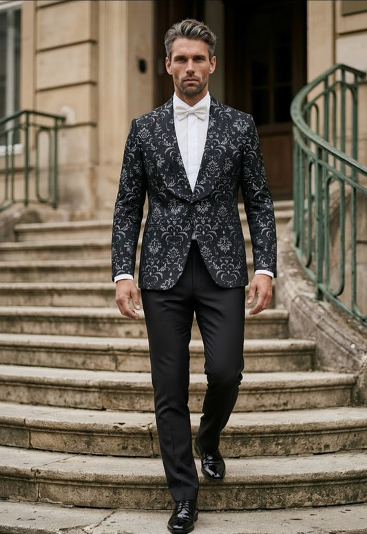 A man wearing a dark patterned tuxedo jacket with a white shirt and bow tie walks down stone stairs in front of a building with green railings.