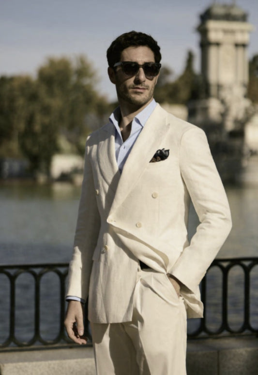 Man in a beige suit and sunglasses standing by a waterfront with trees and a building in the background.