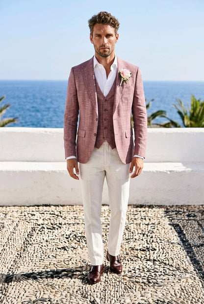 A man wearing a dusty rose three-piece suit with a white shirt, cream trousers, and dark brown dress shoes, standing outdoors on a stone path with the blue ocean and white wall behind him.