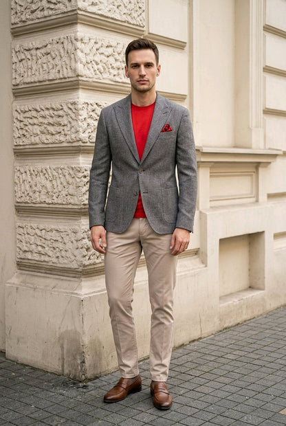 A man wearing a gray textured blazer over a red t-shirt, beige trousers, and brown leather shoes, standing outdoors against a beige stone building.