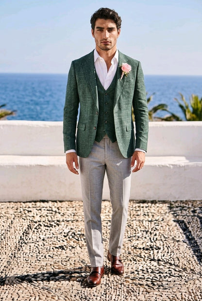 A man wearing a green textured blazer and matching vest over a white shirt, paired with light gray trousers and brown dress shoes, standing outdoors with the ocean and white wall in the background.