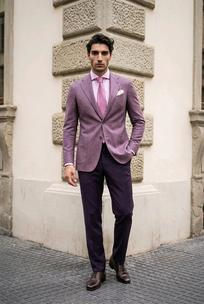 A man wearing a lavender purple blazer, dark purple trousers, a pink patterned shirt, and a matching pink tie, standing outdoors against a light-colored stone building corner.