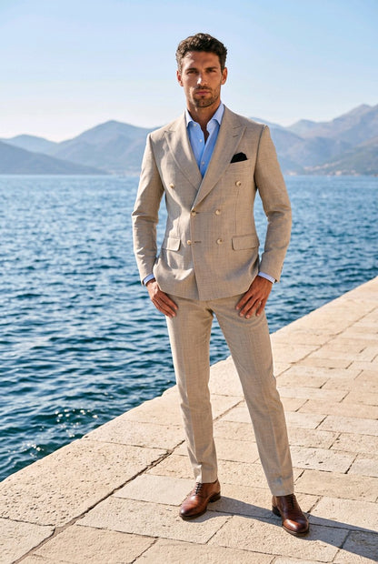 A man wearing a light beige double-breasted suit, a light blue dress shirt, and brown leather shoes stands on a stone pier with a blue sea and mountains in the background.