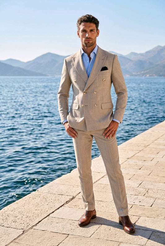 A man wearing a light beige double-breasted suit, a light blue dress shirt, and brown leather shoes stands on a stone pier with a blue sea and mountains in the background.