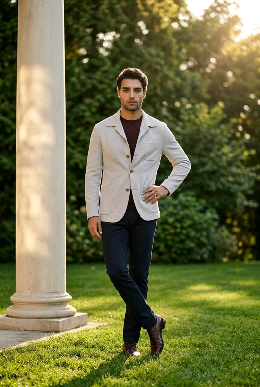 A man wearing a light beige linen-blend blazer over a dark brown turtleneck, navy trousers, and brown shoes, standing outdoors on a green lawn next to a white stone column with warm sunlight filtering through the background trees.