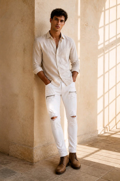 A man wearing a light beige linen button-down shirt, white distressed skinny jeans, and brown leather boots, leaning against a textured beige stone wall in a sunlit room.