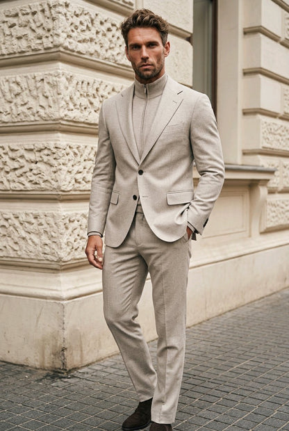 A man wearing a light beige two-piece suit with a mock neck shirt, standing outdoors against a textured cream stone building.