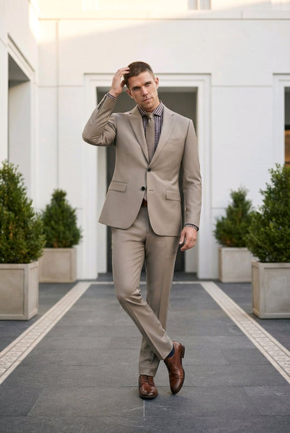 A man wearing a light beige two-piece suit with a patterned purple shirt and brown leather shoes, walking on a paved path in front of a modern white building.