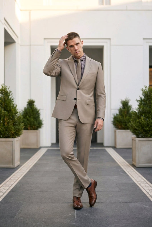 A man wearing a light beige two-piece suit with a patterned purple shirt and brown leather shoes, walking on a paved path in front of a modern white building.