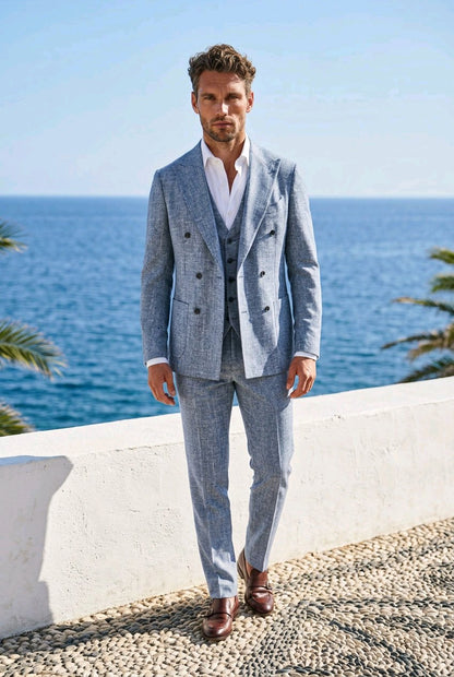 A man wearing a light blue double-breasted suit with a white shirt and brown leather shoes, standing on a pebble path with the blue ocean and sky in the background.