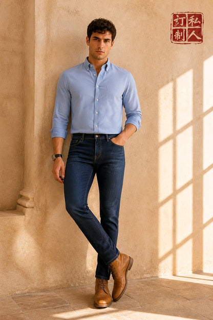 A man wearing a light blue long-sleeve button-down shirt, dark blue jeans, and brown leather boots, standing in a warm, sunlit room with textured beige walls and window shadows. A red Chinese brand logo is visible in the top right corner.