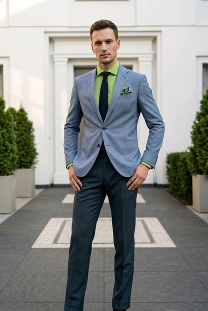 A man wearing a light blue textured blazer, dark navy trousers, a bright green dress shirt, a black tie, and a green pocket square, standing outdoors in front of a white building.