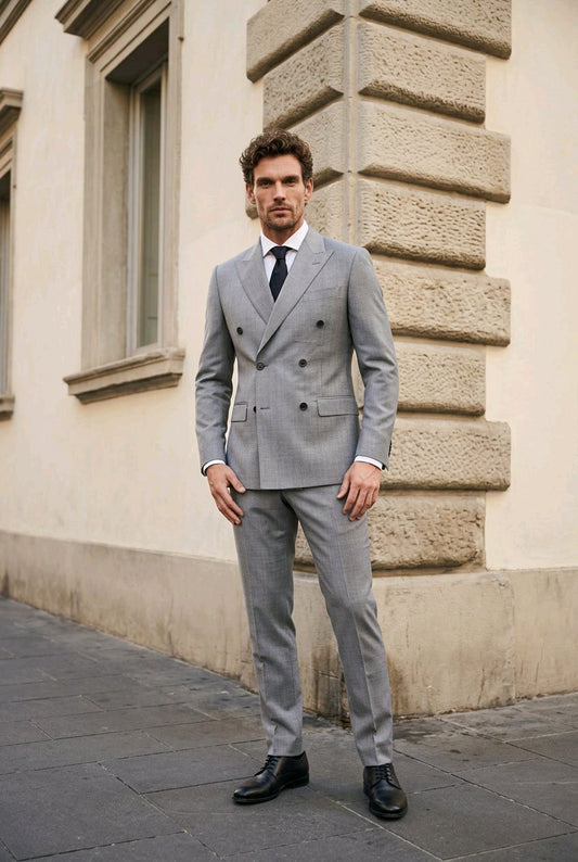 A man wearing a light gray double-breasted suit, white dress shirt, and black tie stands outdoors against a beige stone building corner.