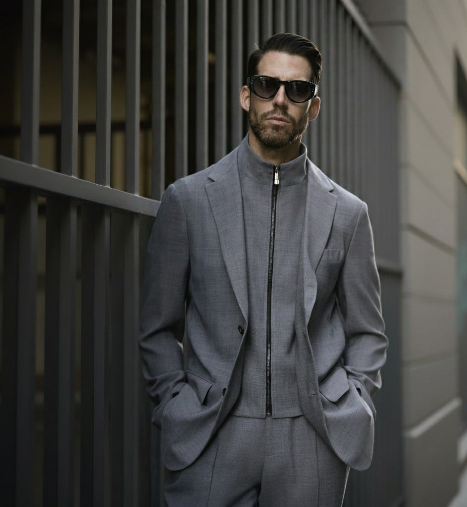 A man wearing a medium grey wool stretch three piece suit with a notched collar and straight flapped pockets, paired with a zippered jacket and trousers. He is standing outdoors with sunglasses on and has a beard.