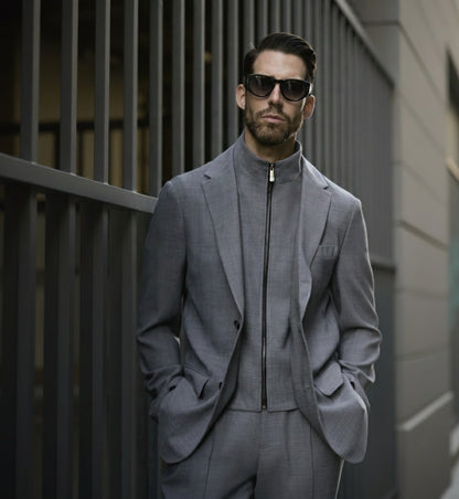 A man wearing a medium grey wool stretch three piece suit with a notched collar and straight flapped pockets, paired with a zippered jacket and trousers. He is standing outdoors with sunglasses on and has a beard.
