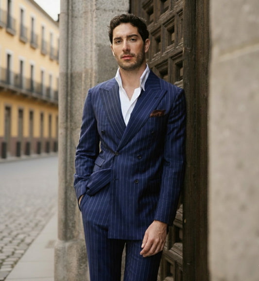 A man wearing a navy blue double-breasted pinstripe suit with a white shirt, standing beside a stone structure.