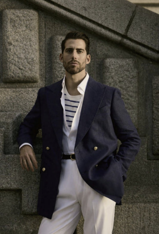 A man wearing a navy blue linen blazer with white cotton-lycra trousers. He is standing in front of a stone wall.