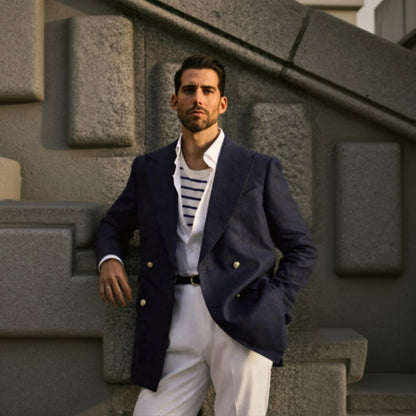 A man wearing a navy blue linen blazer with white cotton-lycra trousers. He is standing in front of a stone wall.
