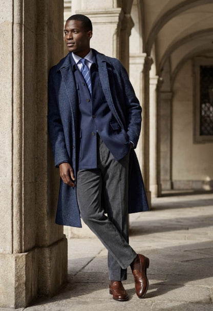 A man wearing a navy blue overcoat, matching navy vest and tie, grey trousers, and brown leather shoes, leaning against a stone column in an outdoor colonnade.