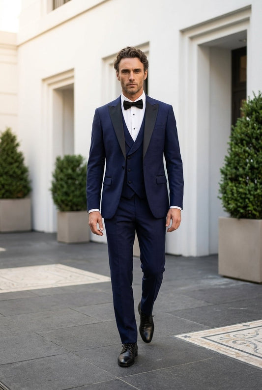A man wearing a navy blue three-piece tuxedo with black satin lapels, a white dress shirt, and a black bow tie walks outdoors in front of a white building.
