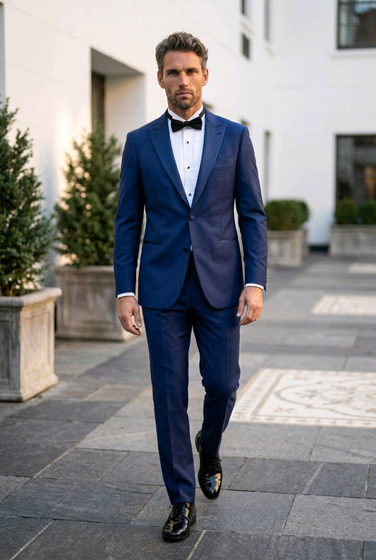 A man wearing a navy blue tuxedo with a white dress shirt, black bow tie, and black dress shoes walks outdoors on a paved walkway in front of a modern white building.