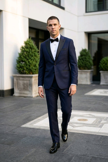 A man wearing a navy blue tuxedo with black satin lapels, a white dress shirt, and a black bow tie, walking outdoors in front of a modern building.