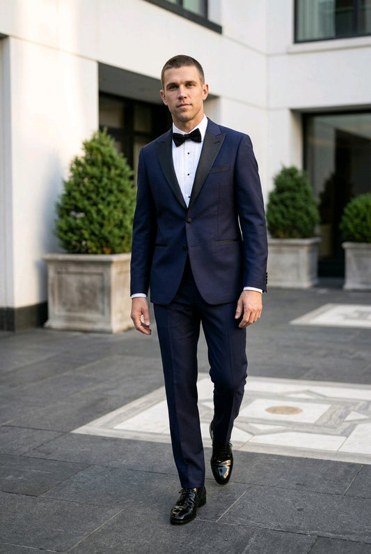 A man wearing a navy blue tuxedo with black satin lapels, a white dress shirt, and a black bow tie, walking outdoors in front of a modern building.