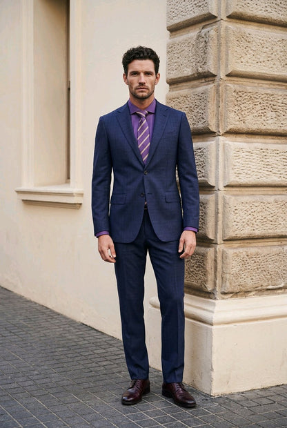 A man wearing a navy blue two-piece suit with a purple patterned tie, standing outdoors against a beige stone building.