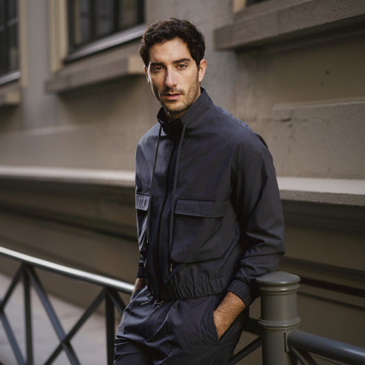 A man wearing a navy two piece suit with a Nehru collar and patched flapped pockets, standing near a railing.