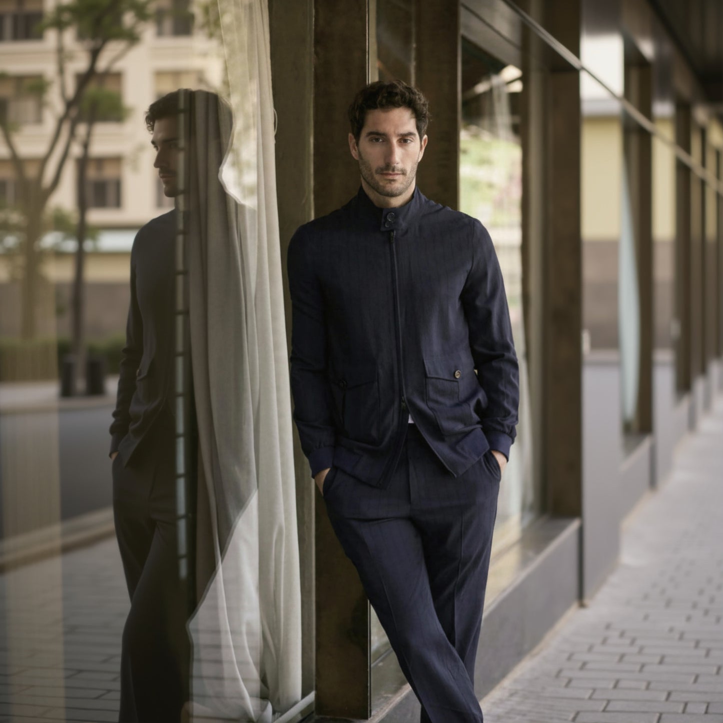 A man wearing a navy wool stripe stretch two piece suit with a Nehru collar and button-up front, standing in front of a glass window.