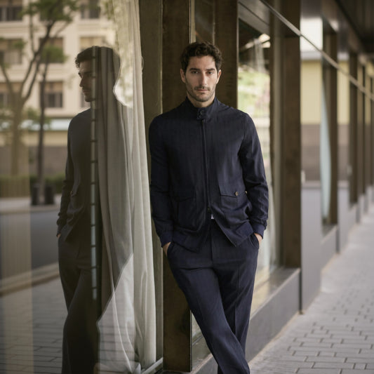 A man wearing a navy wool stripe stretch two piece suit with a Nehru collar and button-up front, standing in front of a glass window.